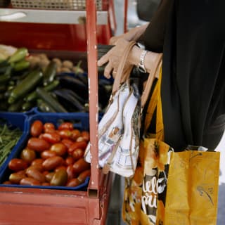 A woman holds her purse and shopping bag at a market stall abundant with plum tomatoes, courgettes and green beans