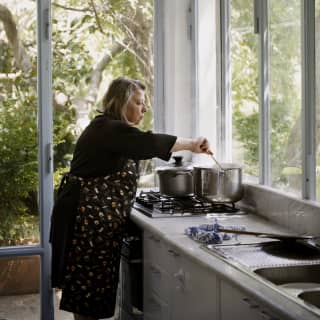 A nonna dressed in black with a patterned apron, stirs a steaming pan on a hob in a rustic kitchen with floor to ceiling windows