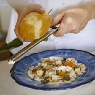 A chef grates the zest of a large lemon onto a salad plate of cubed cheeses, mandarin segments and olives