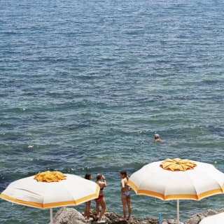 Out to sea, a small boat is shaded by a white canopy. On shore, three girls stand on rocks chatting near flower-like parasols