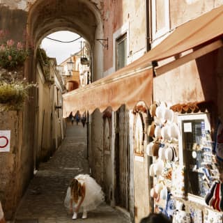 A young girl dressed in a white Confirmation Dress bends to adjust her sock, shaded by an archway on a cobbled Ravello alley