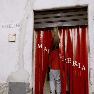 A man parts the ribbons of the red PVC fly curtain across the doorway of a butcher's shop, emblazoned 