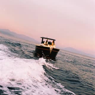 Angled view over the choppy wake of a boat as it carries two guests, seated on the back deck, towards a pink hazy sunset sky.