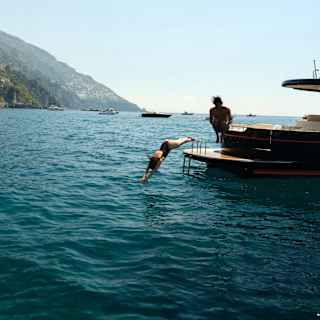 A woman dives from the back deck of a boat as her male friend canon-balls, both captured in mid-air, with an ocean backdrop.
