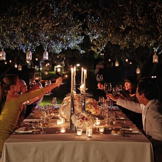A group toasting across a candlelit table under two trees with hanging lanterns above