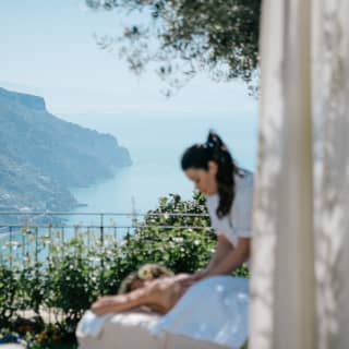 A masseuse with a long dark ponytail, massages her client in the garden shade before cliffs that drop dramatically away