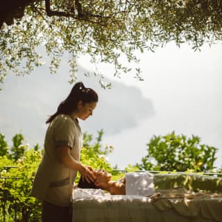 A female spa therapist performs a facial on a guest, who lies on a bed in pure relaxation in the shade of an olive tree.