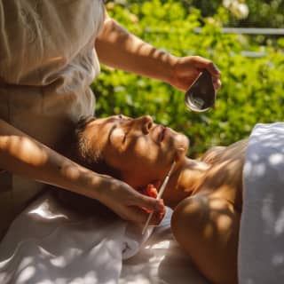 A spa therapist brushes a hydration fluid onto the face of female guest, who lies beneath a cover in dappled shade.