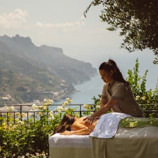 A masseuse works on the back of a woman on an outside treatment bed enjoying physical relaxation and sublime coastal views.