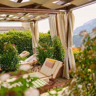 Three lounge chairs with red frames recline beneath a cloth-covered pergola with tied back drapes, viewed through foliage.