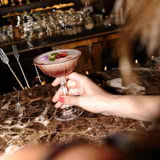 A female guest reaches for a cocktail with pink hues, soft foam and botanical garnish on the marbled bar-top at Bar Taroba.