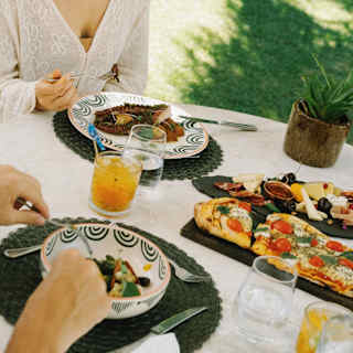 Angled close-up of two guests enjoying light lunch at an al-fresco garden table with pizza, grilled octopus and cheese board.