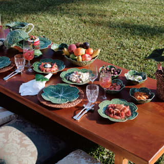 Fruit, charcuterie, salads and smoked salmon are served in green majolica plates and bowls at a picnic table on the lawn.