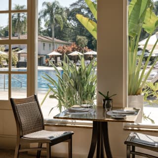 A window table for two enjoys filtered sun and pool views, part obscured by potted strelitzia plants, seen inside Ipê Grill.