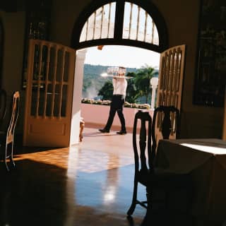 A waiter carries a drinks tray on the balcony of Itaipu restaurant, seen across interior parquet flooring through open doors.