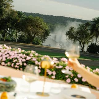 A hand raises a glass of wine at an al-fresco table in soft-focus foreground in an angled shot of the gardens and falls.