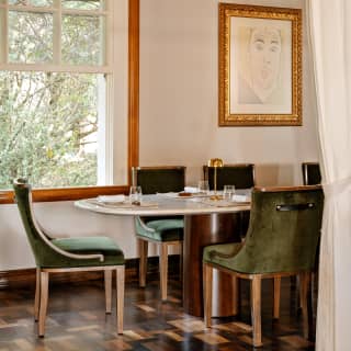 A large window offers garden views to a marble-top dining table with five visible green velvet Art Deco dining chairs.