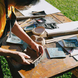 A woman in an apron shapes a clay tile as she attends a ceramics workshop on an outside table covered with tools and clay.