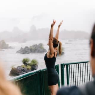 At a viewpoint, a woman in black Lycra and a pony tail faces the Iguassu Falls with arms stretched high, feeling the spray.