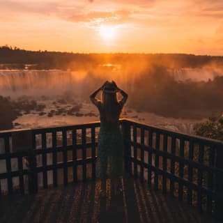 Lady in a green dress and sunhat standing on a platform overlooking the Iguassu Falls in an orange sunrise.