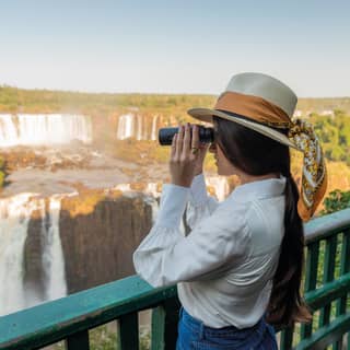 A woman in an elegant sun hat looks through binoculars across the misty cascades of Iguassu Falls at first light