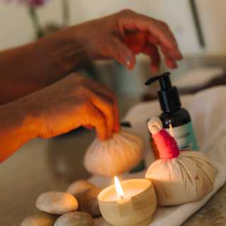 Close-up of a spa table, where a masseuse prepares herbal poultices on a towel, alongside a bottle of oil and massage stones.