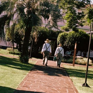 Two gardeners wearing hats and trousers with braces head along a stone tiled garden path towards the hotel, seen from behind.