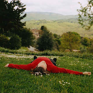 A man in a red jumper lies like a starfish on a hill, feet pointing to the wide Tuscan views, seen from grass above his head.