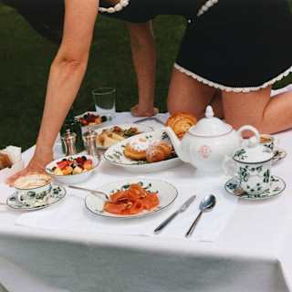 A woman in a black dress with white piping and socks crawls across a breakfast table with pastries, fruit, coffee and salmon.
