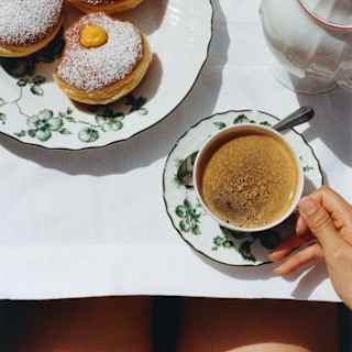 A woman holds the handle of her coffee cup at a table with a plate of three sugar-dusted bombolones, seen from above.