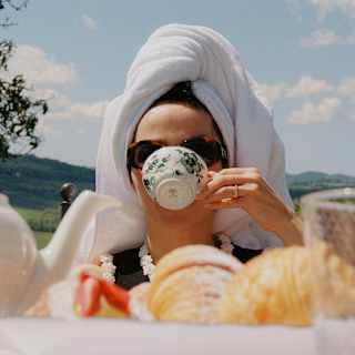 A woman with a towel hair-wrap drinks tea from a porcelain botanical-print cup, seen at eye-level over a table of pastries.