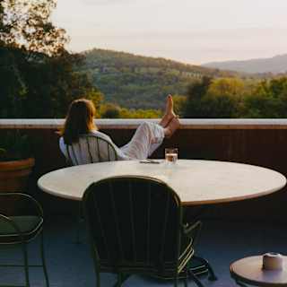 A dark-haired woman in white sits a terrace table, bare feet up on the wall, gazing at gold-tinted hills, seen from behind.