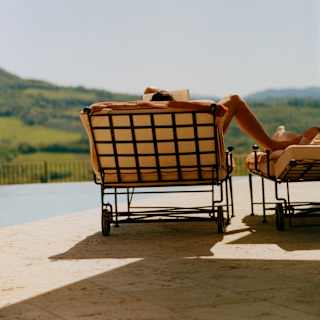 A man reads on a poolside lounger, one leg extended to rest on an adjacent chair, seen from behind in an image of serenity.