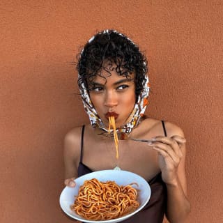 A young woman in a patterned Jackie O headscarf stands beside a deep sienna wall eating a plate of spaghetti al ragù