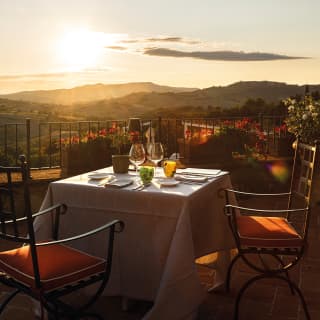 A dining table for two nestles in the corner of a terrace overlooking the rolling Tuscan hills, painted gold by evening sun.
