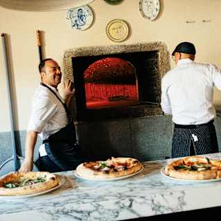 Two chefs in white with blue aprons stand by the red-hot wood-fired oven, seen over a counter with three pizzas ready to go.