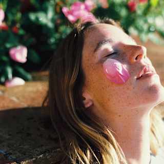 A woman with shoulder-length blonde hair and freckles leans back against a stone wall with a pink rose petal on her cheek.