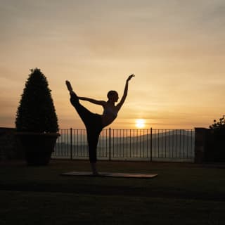 A young woman stands on a yoga mat in a perfectly balanced dancer pose as the sun slowly climbs into dawn behind her