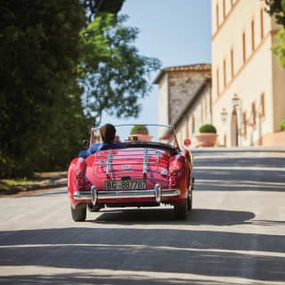 Red vintage soft-top car driving along a tree-lined driveway towards a Tuscan castle
