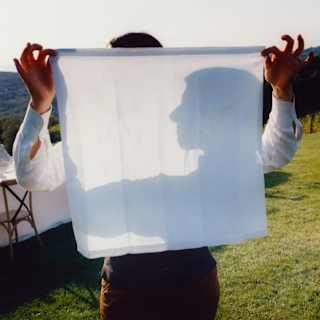 A man holds up a white napkin which shows the shadow of his face in profile, seen at a celebration table in the garden.
