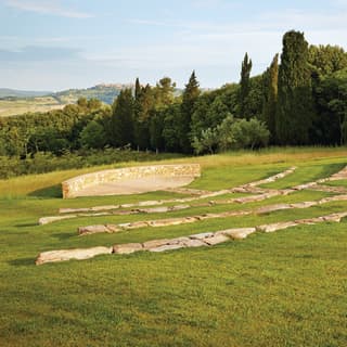 A gently sloping lawn surrounds an ancient Etruscan amphitheatre. In the distance tall trees and sloping hills