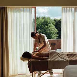 In a mellow room, a spa therapist massages the bare back of a guest lying face down on a bed and half-covered with a sheet.