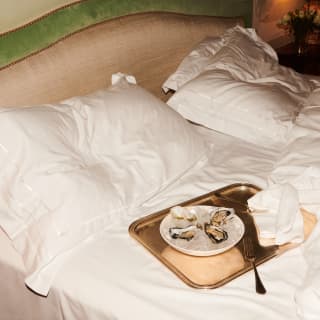 Four fresh oysters lie in a bowl of ice on a golden tray with two forks. The tray sits ready on a recently vacated bed