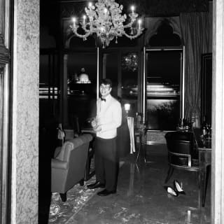 A waiter in a white jacket smiles to the camera as he serves wine in a private lounge below a Murano glass chandelier