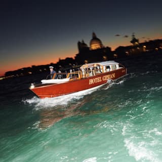 The Hotel Cipriani private launch cuts a bow wave through the green water at dusk. In the background, the lights of St Marks