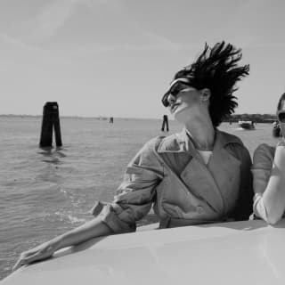 A chic young woman in a classic trench coat and sunglasses allows the wind to catch her hair aboard a boat on the lagoon