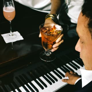 A man plays a polished black grand piano. Lying on it, a woman hands him an iced cocktail. In front of her is a fresh Bellini