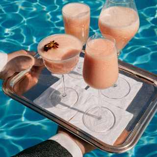 Close-up on a tray of Peach Bellini cocktails served in four different-shaped glasses with a shimmering pool-water backdrop.
