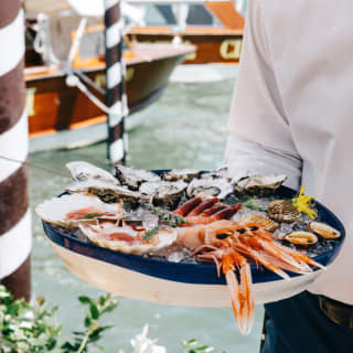 A waiter carries an iced seafood platter. Behind him the Hotel Cipriani’s private launch is moored to one of the paline de casade