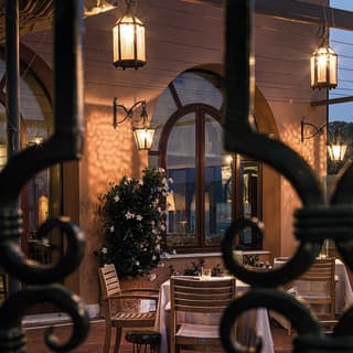 Glass lanterns throw golden patterns onto the pink hotel walls at dusk. The restaurant terrace appears through ornate ironwork